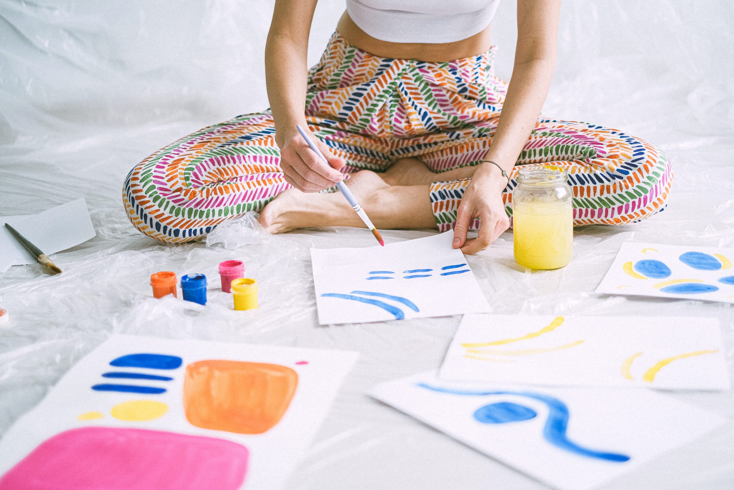 Kobieta w kolorowych spodniach siedząca na podłodze i malująca obraz farbami