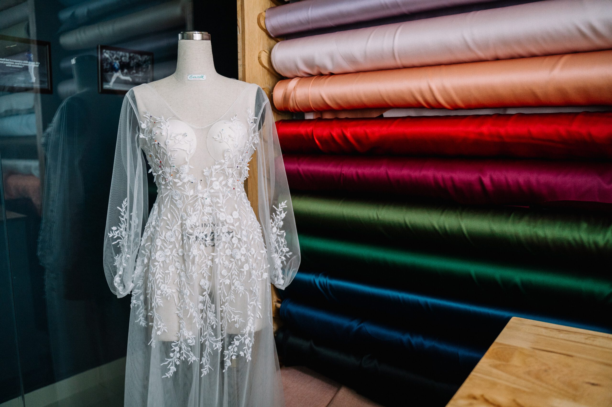 Suknia ślubna na manekinie na tle kolorowych tkanin z poliestru nawiniętych na bale w pracowni krawcowej